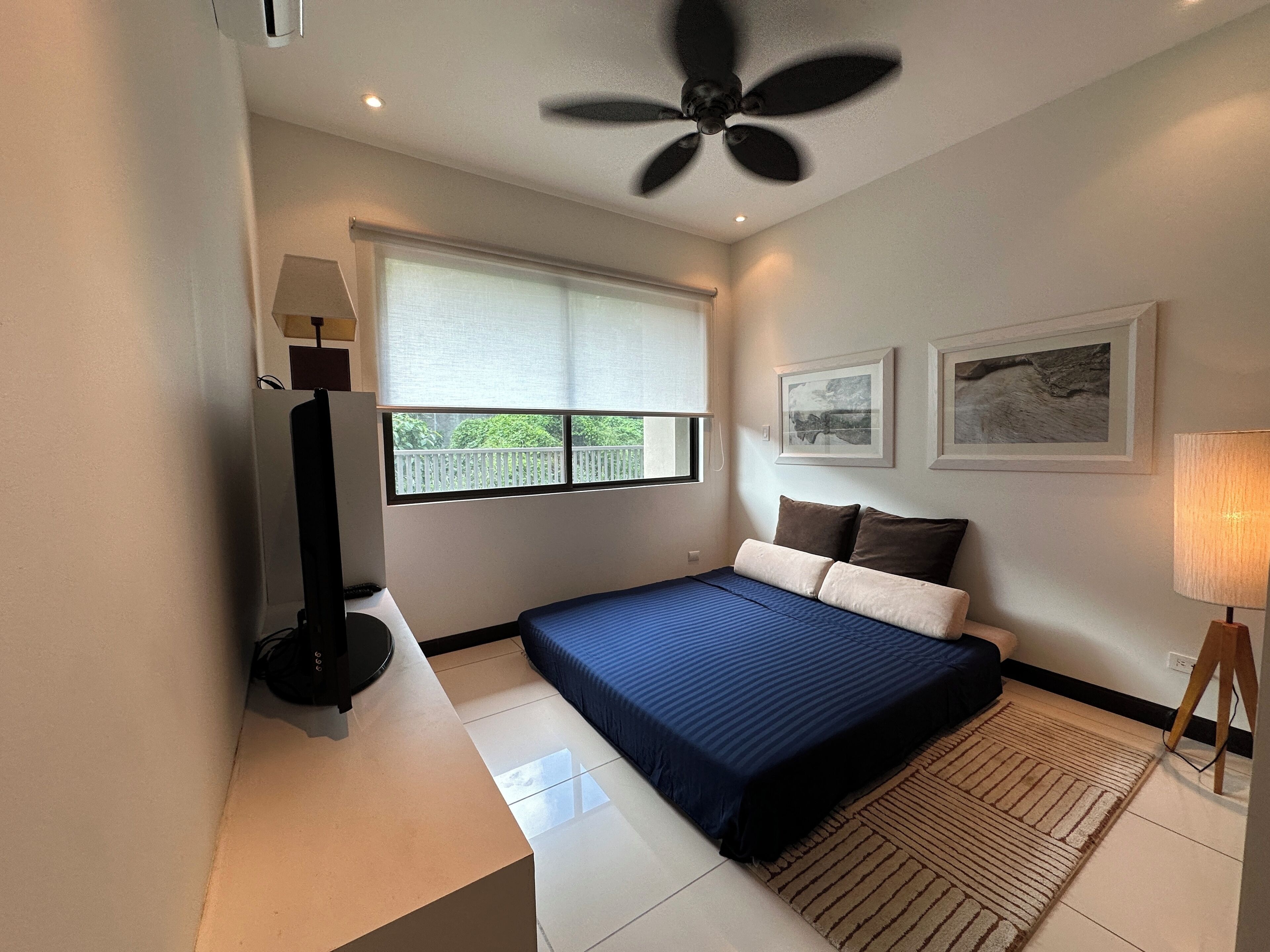 Un dormitorio con una cama, ventilador de techo, ventana con persianas, mesita de noche y una lámpara.