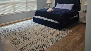 Une chambre à coucher comprenant un lit, deux tables de chevet, un ventilateur de plafond, une fenêtre avec des stores et un tapis à motifs.