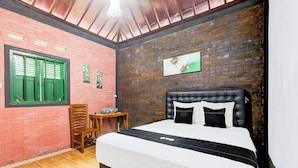 A bedroom with a brick wall, a bed with black and white bedding, a wooden chair, a small table, and a window with green shutters. - Capital O 94018 Joglo Eyang Niek Turi Syariah (Kaliurang)