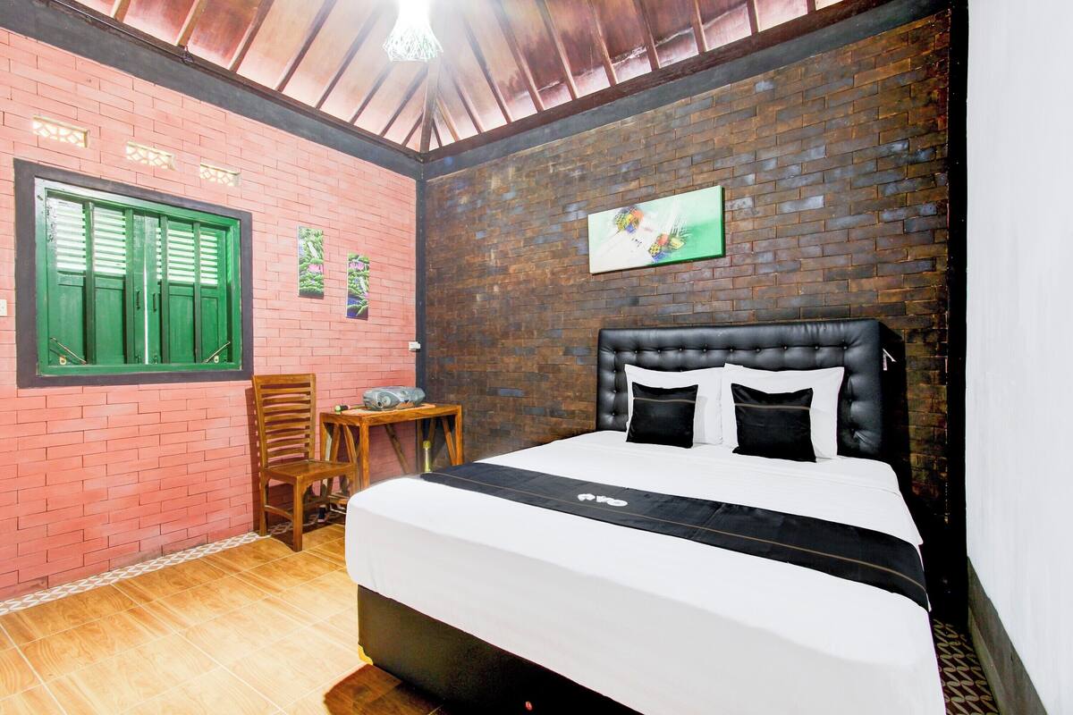 Quarto com parede de tijolos, cama com roupa de cama preta e branca, uma cadeira de madeira, uma mesa pequena e uma janela com persianas verdes.