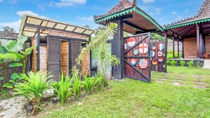 A traditional house with a thatched roof, a wooden gate, and a garden with tropical plants. - Capital O 94018 Joglo Eyang Niek Turi Syariah (Kaliurang)