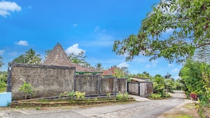 A traditional house with a tiled roof, surrounded by a stone wall and lush greenery. - Capital O 94018 Joglo Eyang Niek Turi Syariah (Kaliurang)