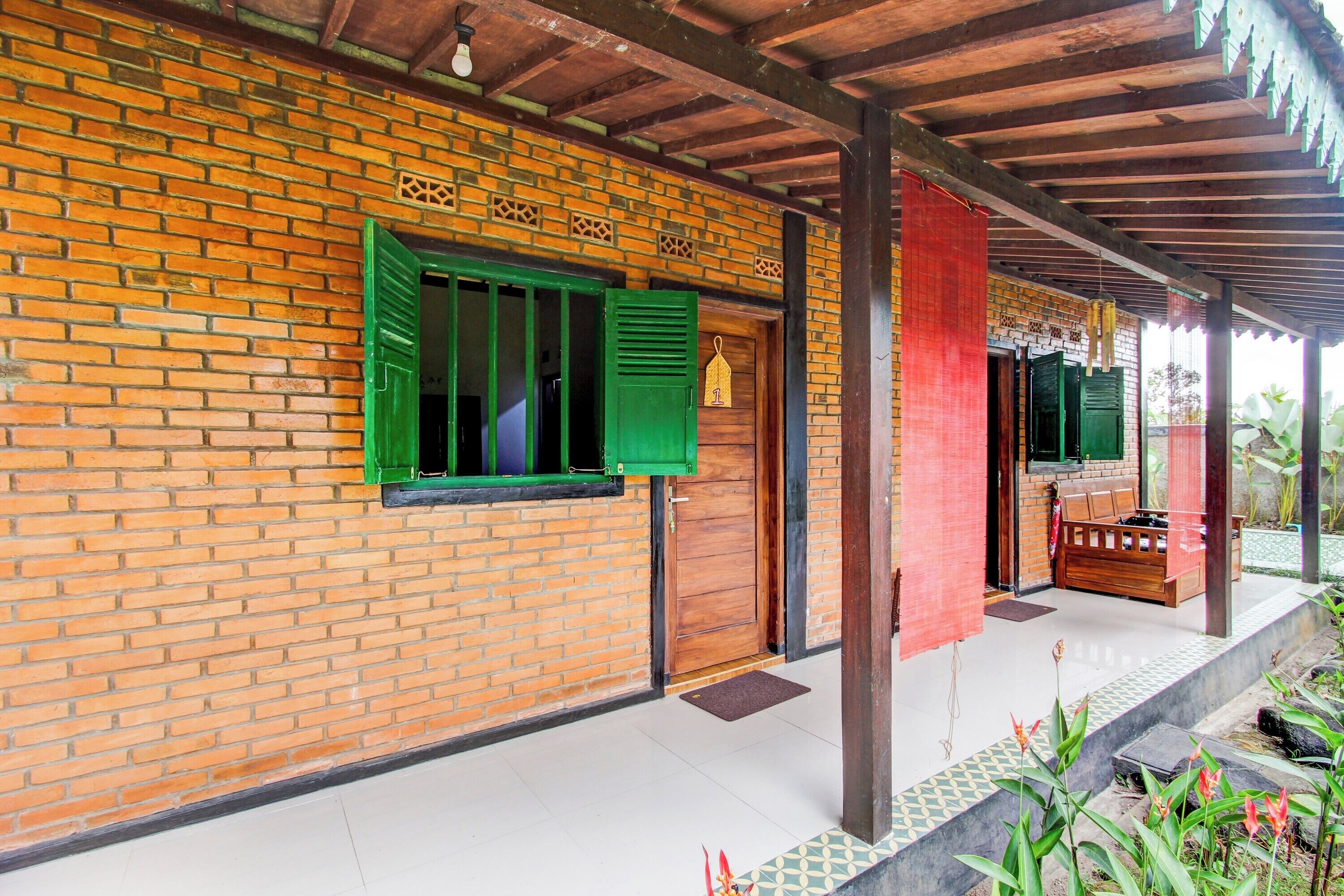 A brick building with green shutters, a wooden door, and a covered porch area with a bench.