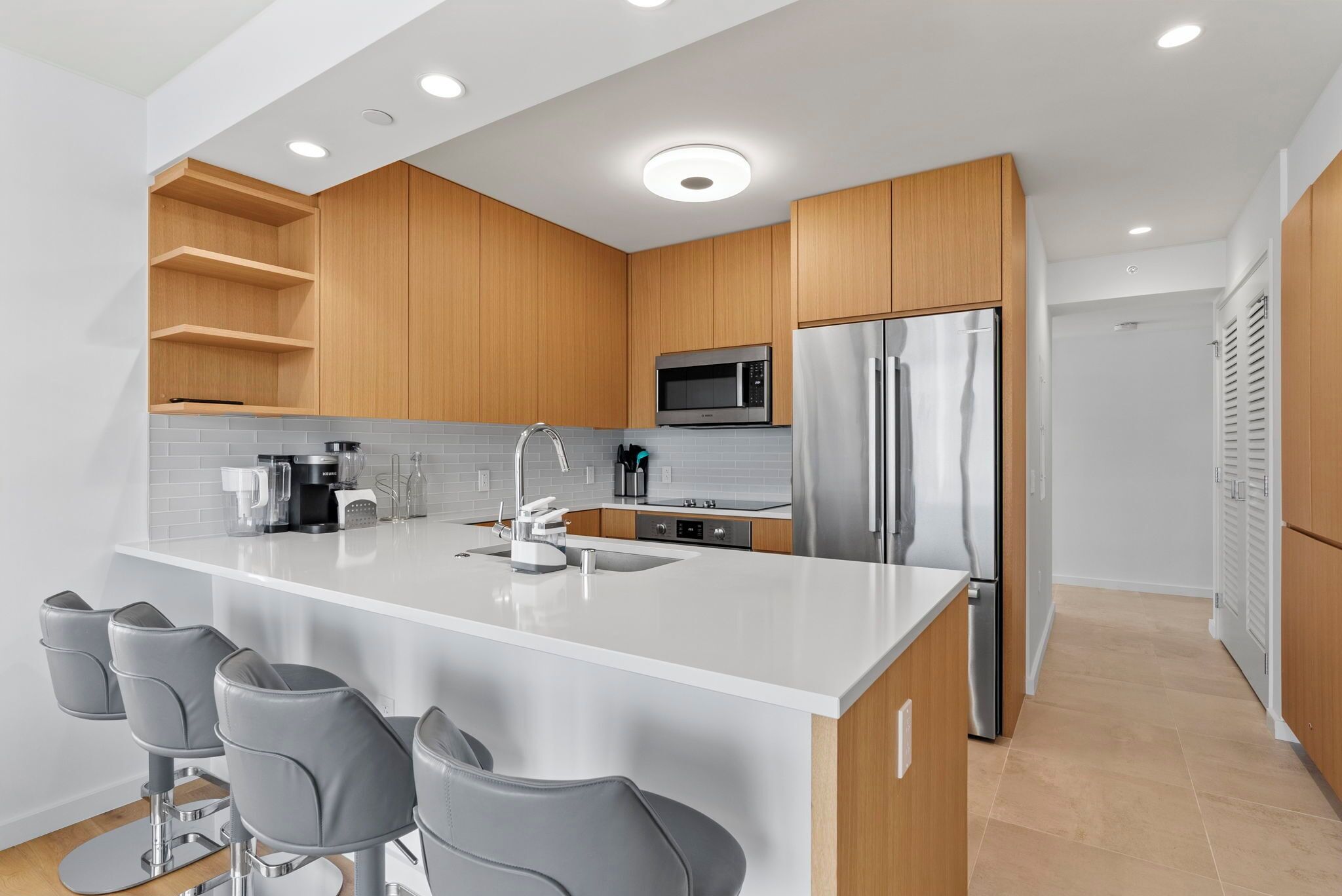 A modern kitchen with wooden cabinets, a stainless steel refrigerator, a microwave, and a bar area with grey stools.