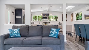 A modern living room with a grey sofa, two blue pillows, a dining area with a table and chairs, and a kitchen in the background.