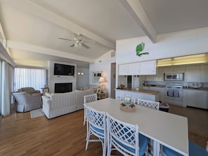Ein modernes Wohnzimmer mit Kamin, einem Essbereich mit weißen Stühlen, einer Küche mit weißen Schränken und einem Deckenventilator.