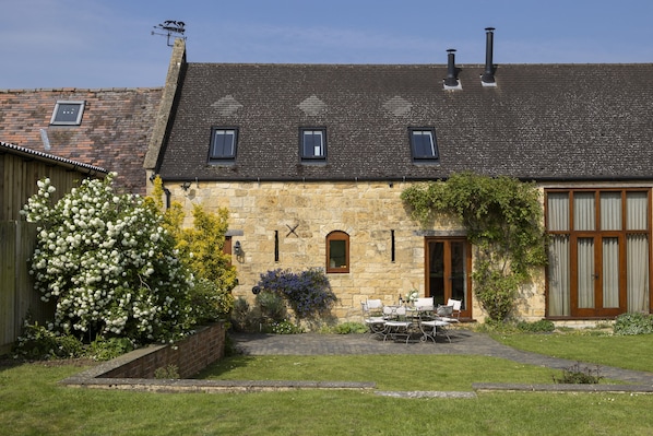 A stone building with a sloped roof, a brick wall, and a small garden with a table and chairs.