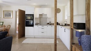 Une cuisine moderne avec des armoires blanches, un four encastré et un four à micro-ondes. On y trouve une structure en poutres apparentes, un coin repas avec des chaises et un tapis blanc sur le sol.