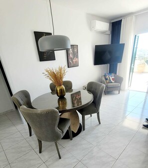 A modern living room with a round glass table, grey upholstered chairs, a black floor lamp, a flat-screen TV, and a large window with curtains. - PJ's Apartment ,Chlorakas, Paphos. 2 Bedrooms with a large south facing veranda. (Chloraka)