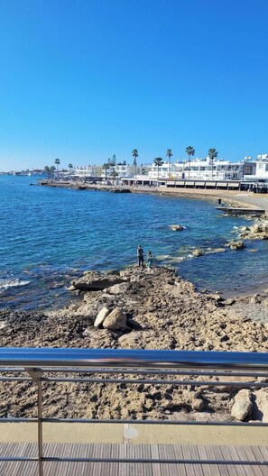 A rocky shoreline with clear blue water, a wooden boardwalk, and a row of buildings in the background. - PJ's Apartment ,Chlorakas, Paphos. 2 Bedrooms with a large south facing veranda. (Chloraka)
