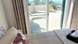 Une chambre à coucher avec un lit, une fenêtre avec des rideaux et une vue sur un balcon agrémenté d’une chaise et d’une plante.