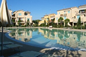 A pool area with lounge chairs and umbrellas, surrounded by multi-story buildings with balconies. - Village of Flowers (Lido di Jesolo)