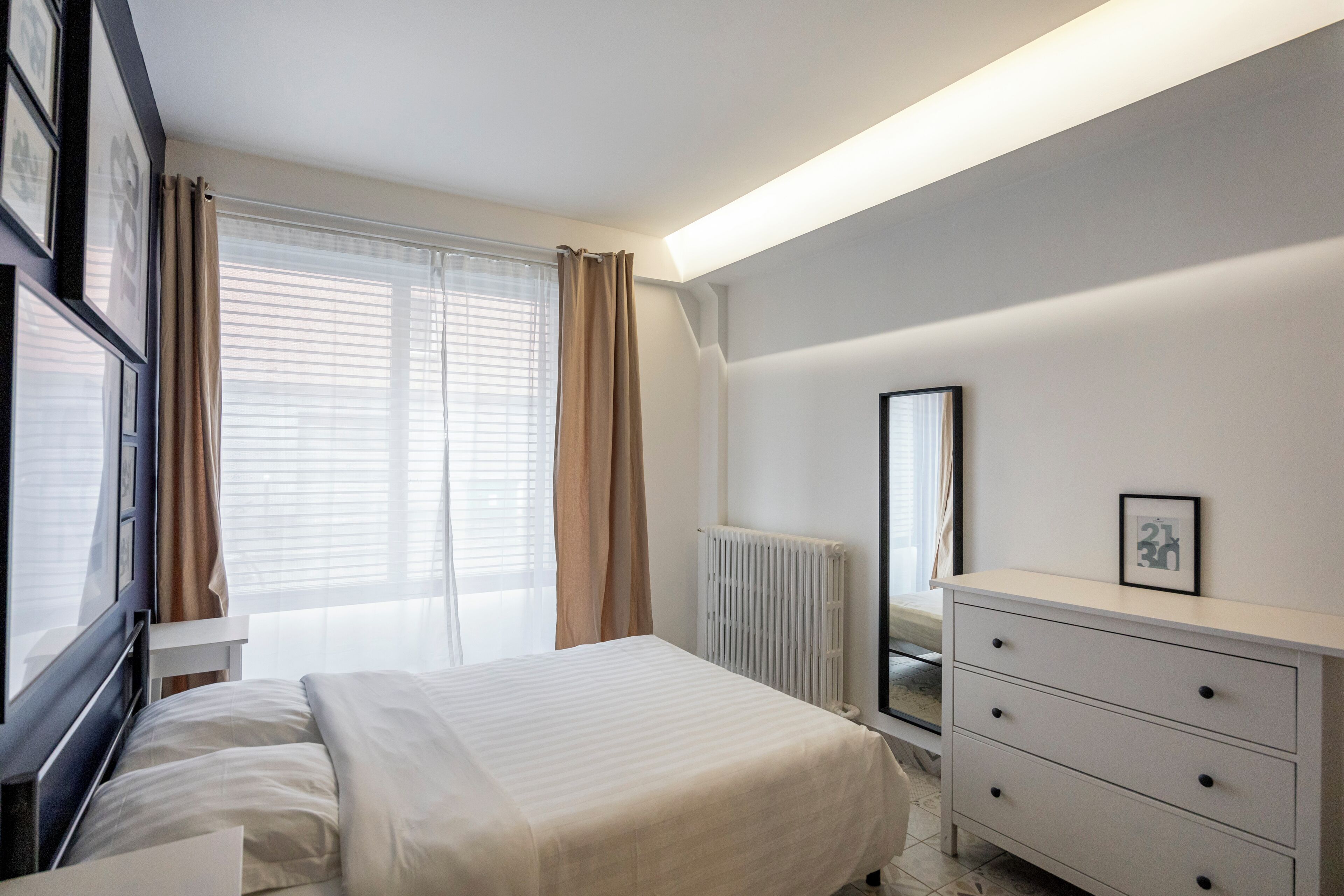 Un dormitorio con cama, cómoda, radiador y ventana con cortinas.