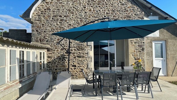 A stone building with a patio area featuring a table and chairs, a lounge chair, and a large umbrella. - Superbe Maison Moderne Entièrement Rénovée Trégomeur Proche Mer - Chez Alphonse (Trégomeur)