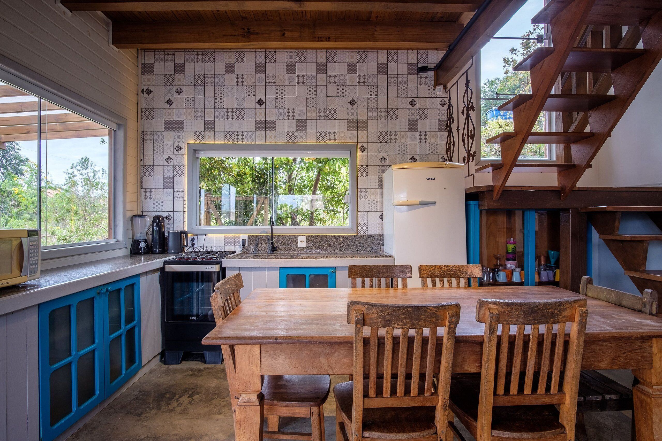 A kitchen with a wooden table and chairs, a refrigerator, and a window with a view of greenery.