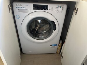 A compact, white Candy Smart washing machine with a digital control panel, located in a small laundry room with a wooden floor.