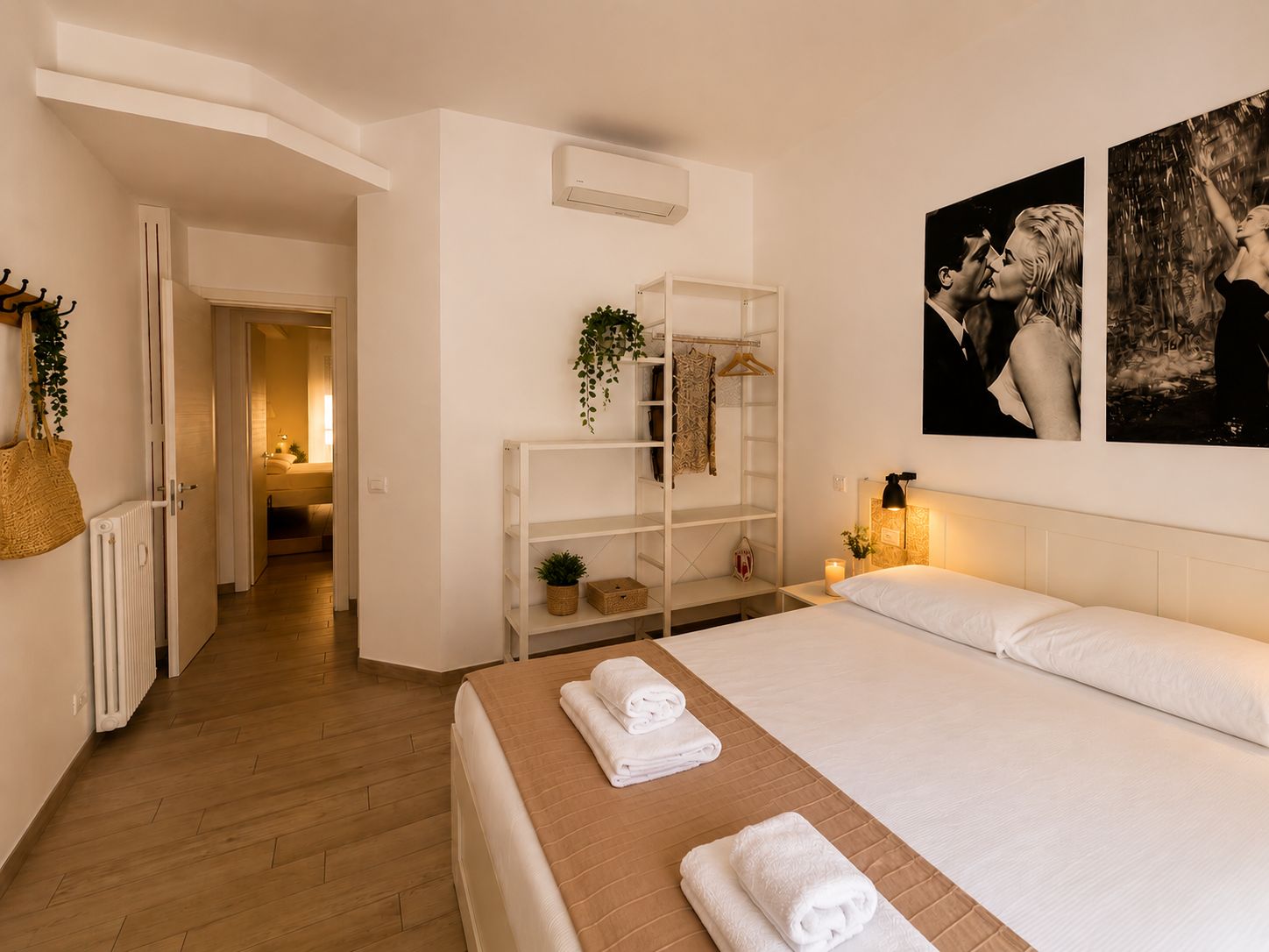 A hotel room with a bed, a bedside table with a lamp, a shelf with decorative items, and a hallway leading to another room.