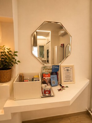 Zona de recepción de hotel con mostrador blanco, un espejo grande, una planta en maceta y folletos informativos.