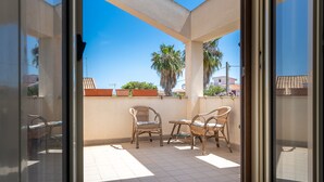 A balcony with two wicker chairs, a tiled floor, and a view of palm trees and buildings.