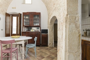 A kitchen with a white tiled floor, a wooden cabinet with glass doors, a television, and a dining table with chairs.