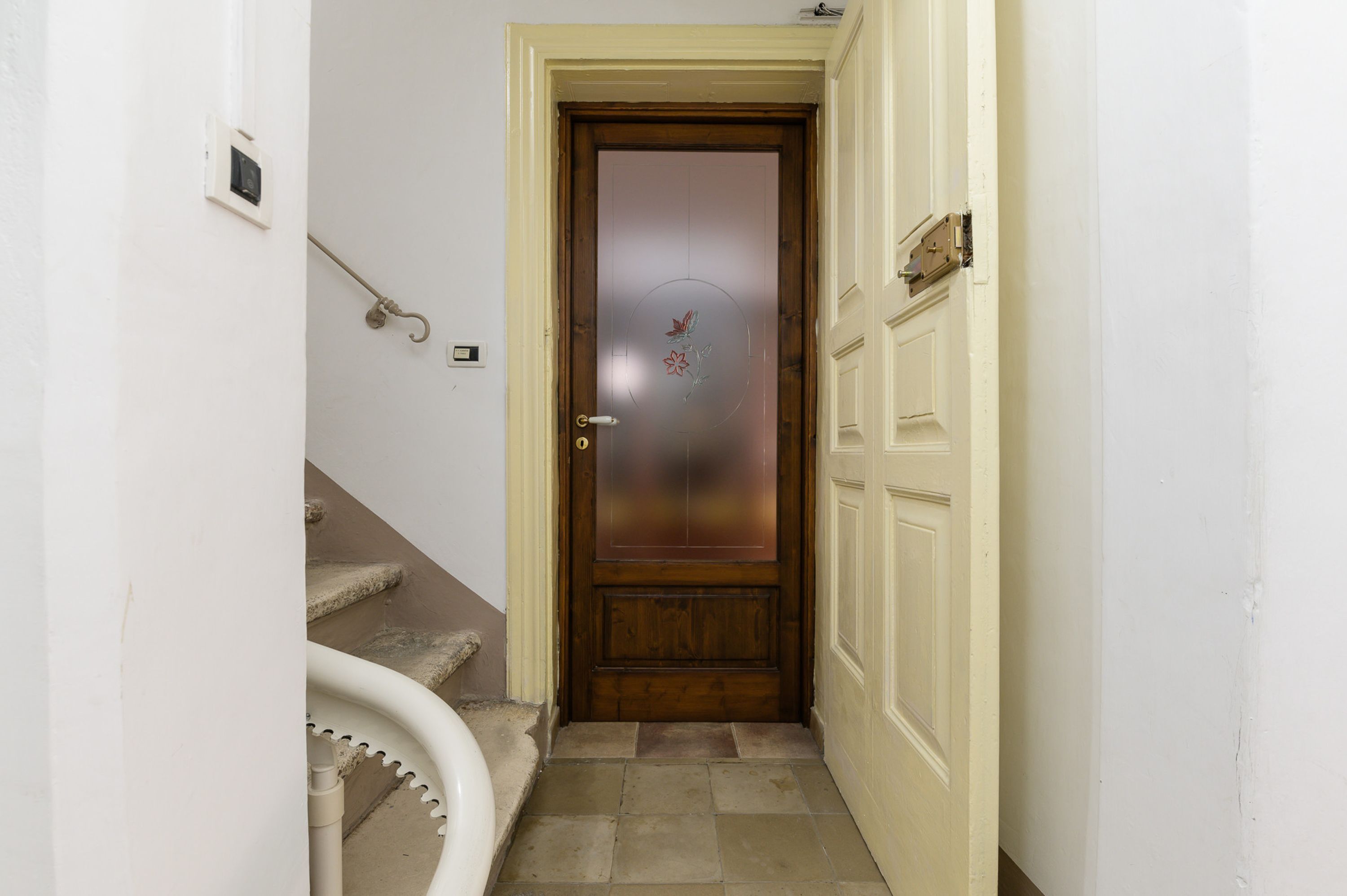 A narrow hallway with a wooden door featuring a frosted glass panel and a decorative emblem. A white wall-mounted light switch and a white pipe are visible on the left.