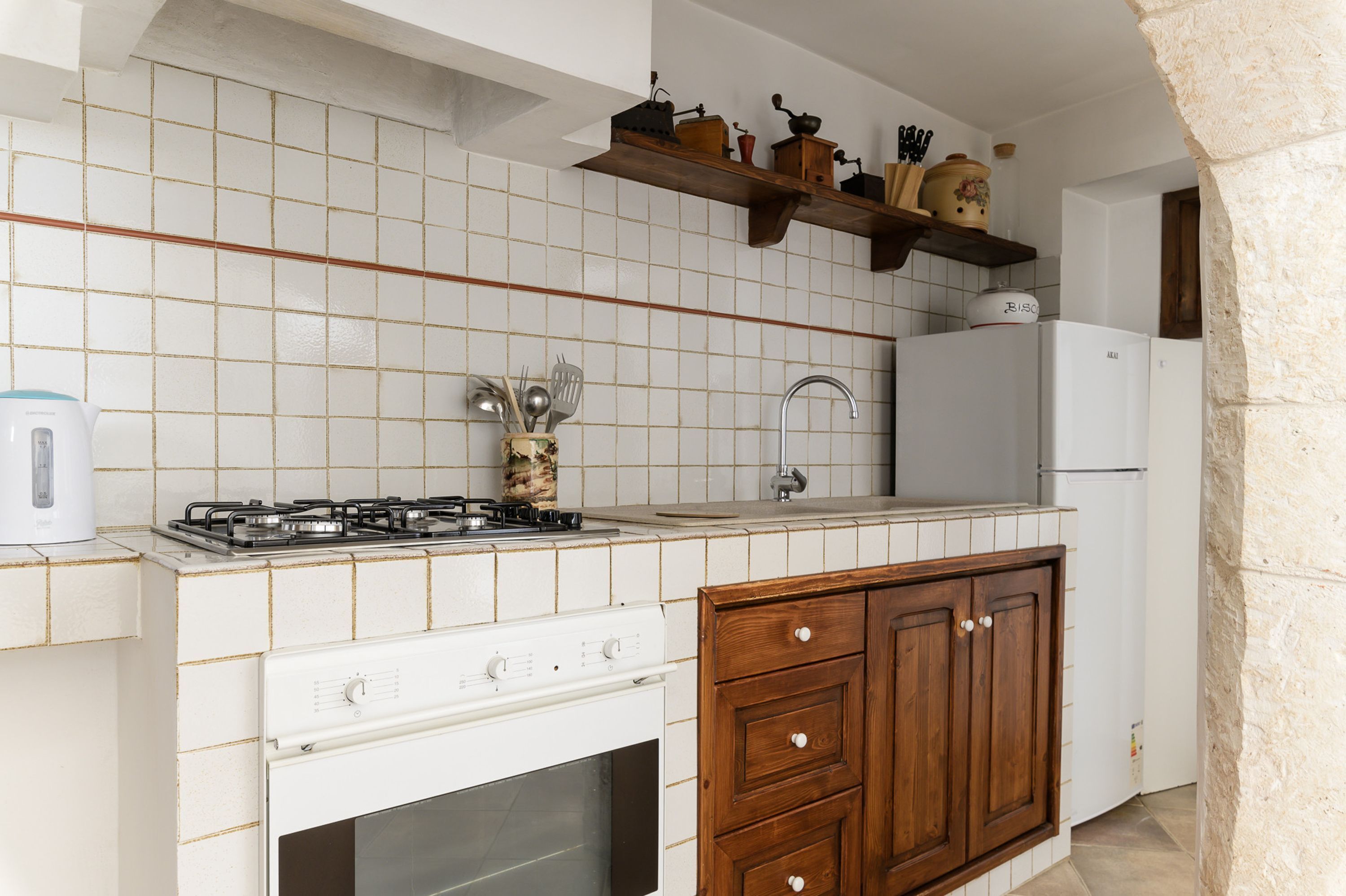 A kitchen with white tiled walls, a gas stove, a sink, wooden cabinets, and a white refrigerator.