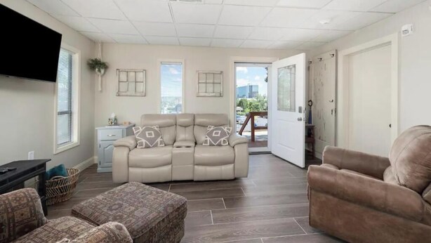 A living room with a beige sofa, a brown armchair, and patterned ottoman. There is a television on the left, a small table with a plant, and a window with blinds. - Branson Downtown Trolley House (Branson)