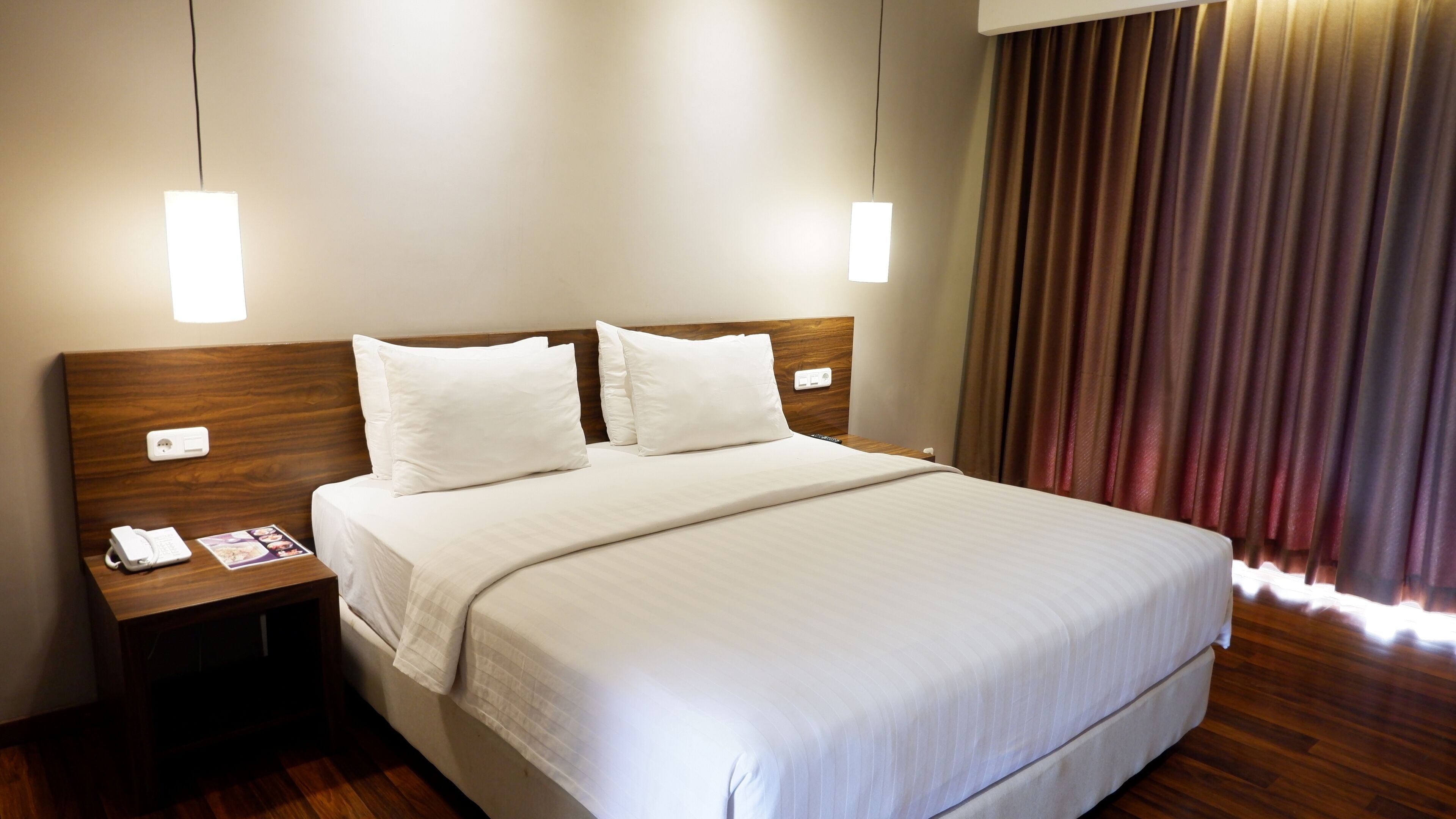 A hotel room with a wooden headboard, two white pillows, a neatly made bed, a bedside table with a phone and a magazine, and a window with curtains.