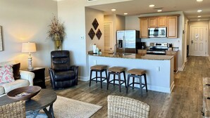 A modern kitchen with a central island, dark wooden flooring, and a seating area with a sofa and chairs. - FREE Daily Golf! Reflections 506 Amazing Views, Heated Resort Pool & Hot Tub! (Panama City)