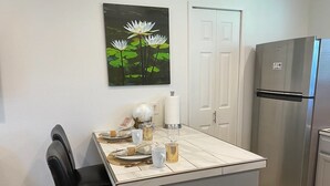 Une cuisine avec une table à manger pour quatre personnes, un réfrigérateur et un tableau représentant des fleurs blanches accroché au mur.