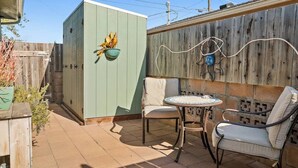Une terrasse avec deux chaises et une table, une clôture en bois et un coffret électrique vert agrémenté d’un pot de fleurs.