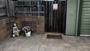 Une clôture en bois, une statue de Bouddha, une plante en pot et un paillasson avec l’inscription « BIENVENUE » à l’entrée d’une propriété.