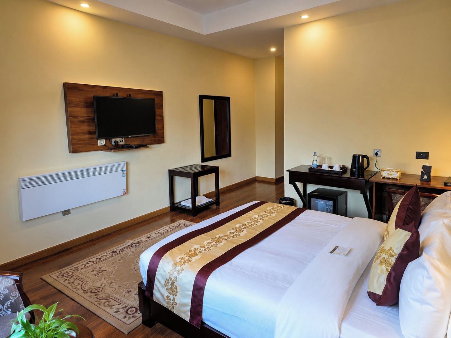 A hotel room with a bed, a desk, a chair, a television, a mirror, and a radiator.