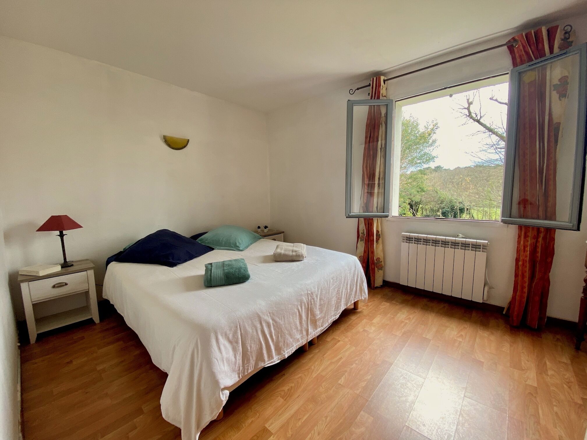A bedroom with a bed, a nightstand, a window with curtains, and a radiator.