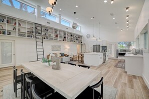 Et rummeligt køkken med en central ø, et spiseområde med bord og stole, og en stor reol fyldt med bøger.