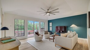 A modern living room with a ceiling fan, a TV, and a large window with a view of the outdoors. - 3-Acre Luxury 5BR Ranch, Movie Theater, 2 Kitchens, Near COTA Airport! Sleeps 14 (Austin)