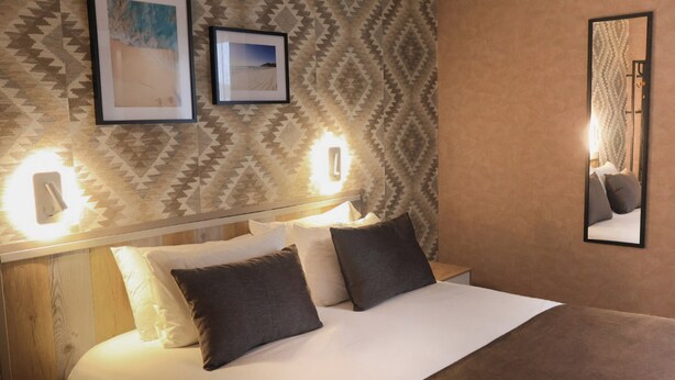 Un lit bien fait, avec une tête de lit, deux tableaux encadrés accrochés au mur et un miroir.