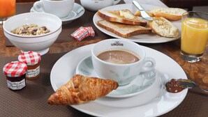 Une table dressée pour le petit-déjeuner avec un bol de céréales, un croissant, une tasse de café, des toasts, de la confiture et un verre de jus d’orange.