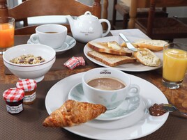 Uma mesa de café da manhã com tigela de cereal, croissant, xícara de café, torradas, geleia e um copo de suco de laranja.