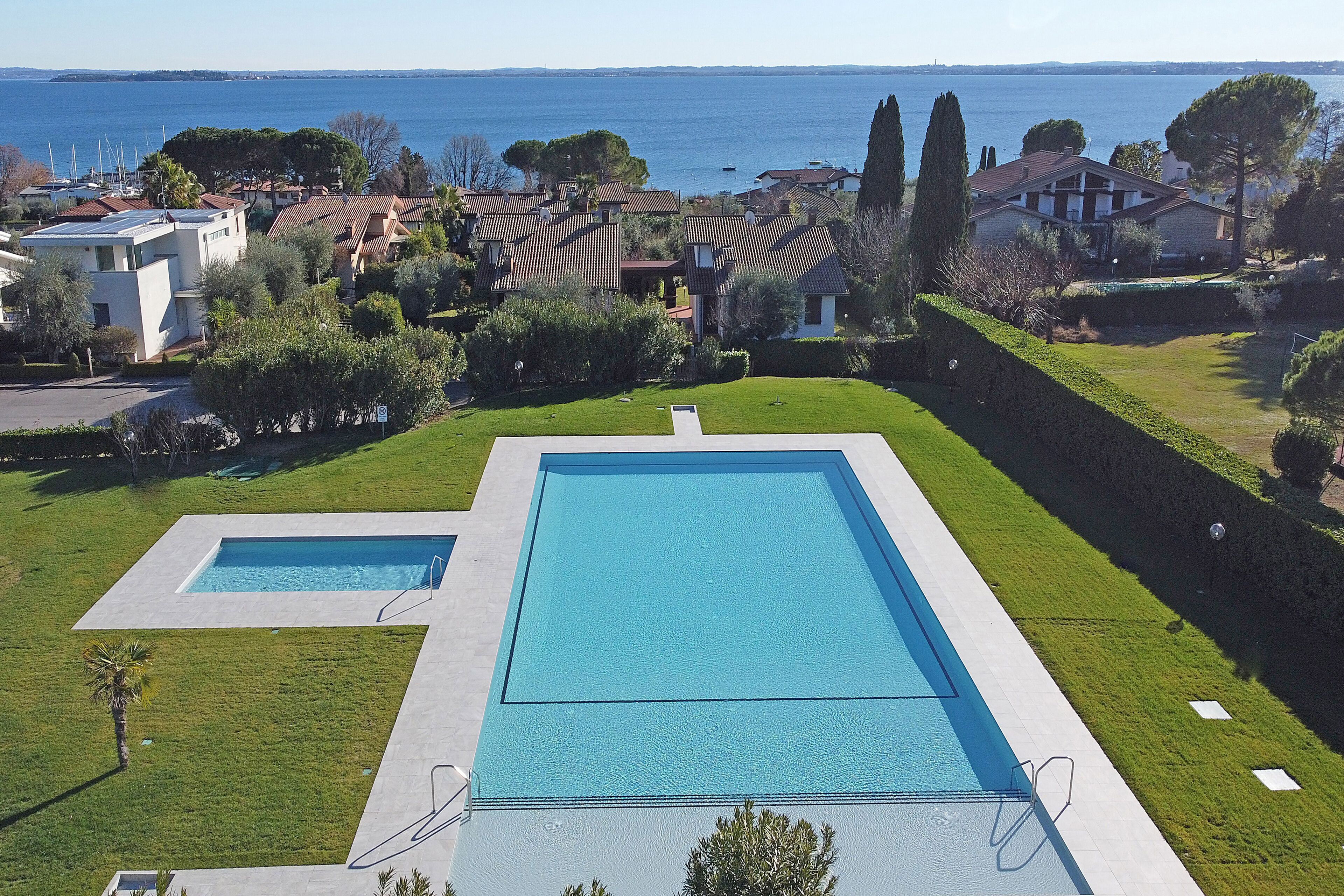 A residential area with a large rectangular swimming pool, surrounded by well-maintained lawns and a view of the sea.