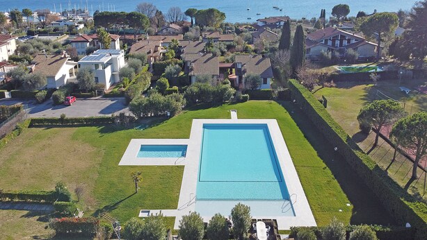 A residential area with a large rectangular swimming pool, surrounded by well-maintained lawns and trees. - Residence Moniga Porto 1-08 by Wonderful Italy (Moniga del Garda)