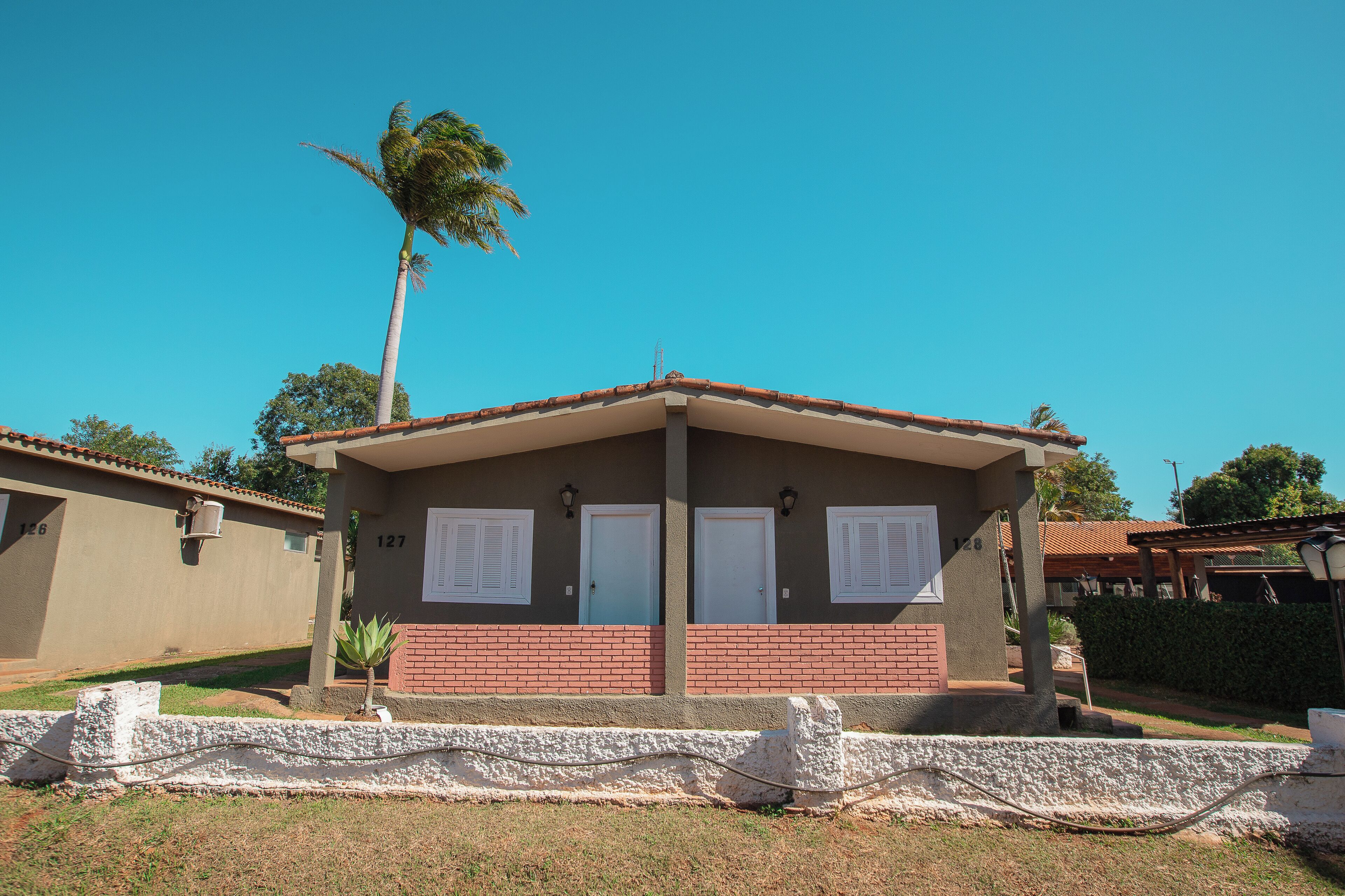 A single-story building with a red-tiled roof, two doors, and a small garden area with a palm tree.