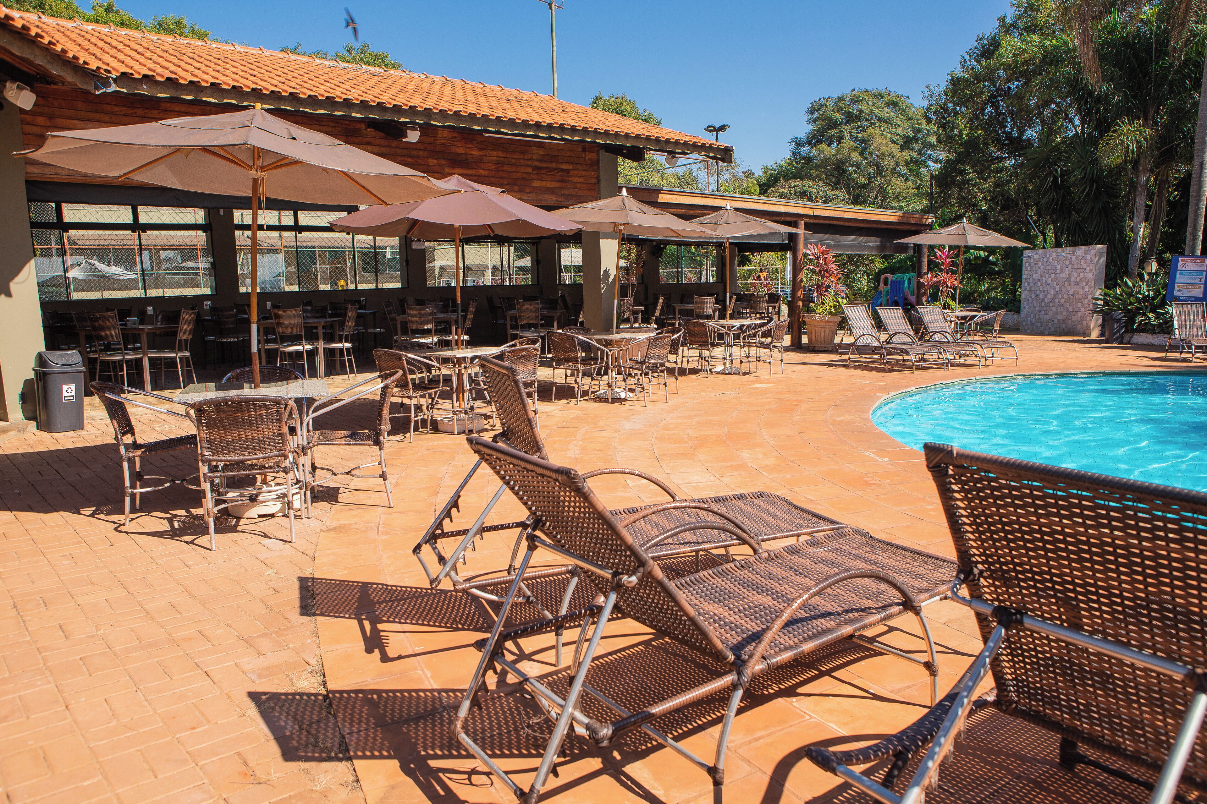 Ein Poolbereich mit Liegestühlen und Sonnenschirmen, einer gefliesten Terrasse und einem Gebäude mit großen Fenstern.