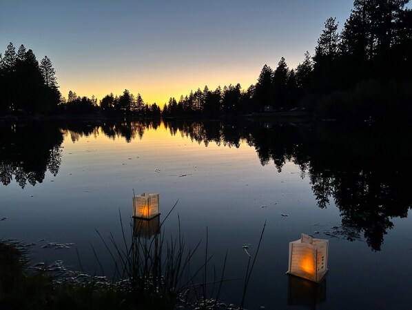 Un lago tranquilo al atardecer con dos faroles iluminados en primer plano y árboles reflejados en el agua.