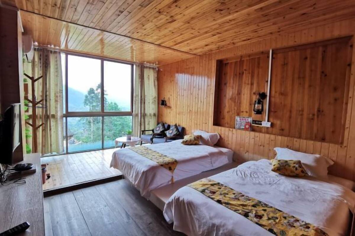 A wooden cabin room with two beds, a TV, and a large window overlooking a forested area.