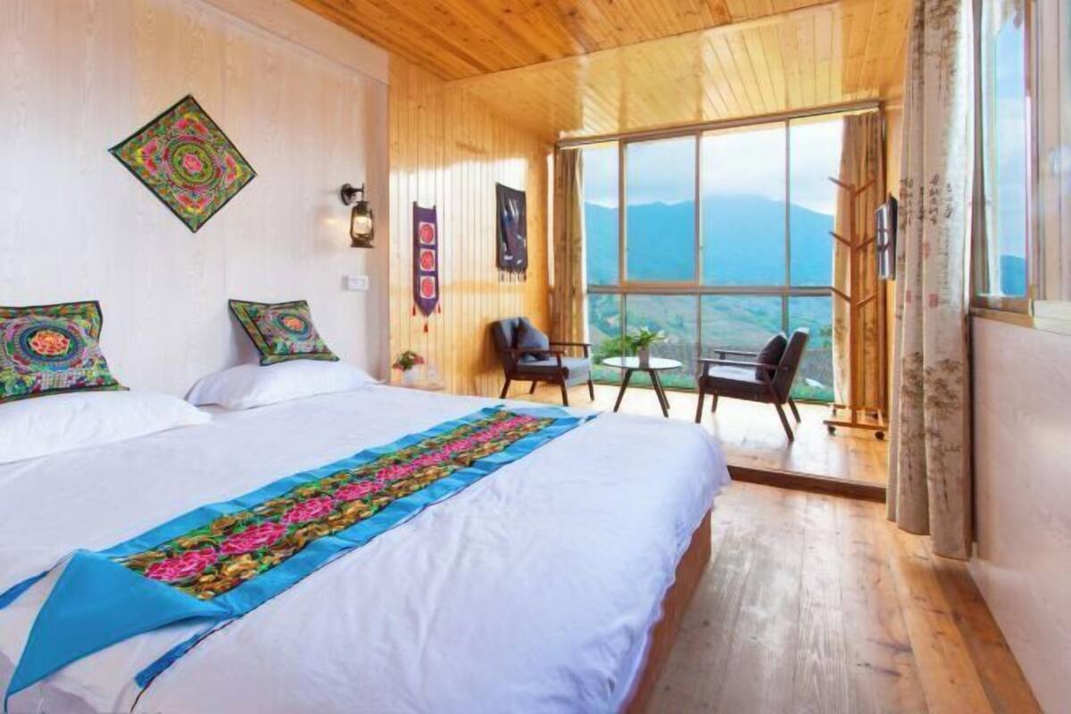 A room with a large bed, a small table, and a chair, with a view of mountains through a large window.