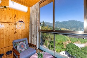 A wooden interior with a large window, a chair with a patterned cushion, and a small table with a plant.