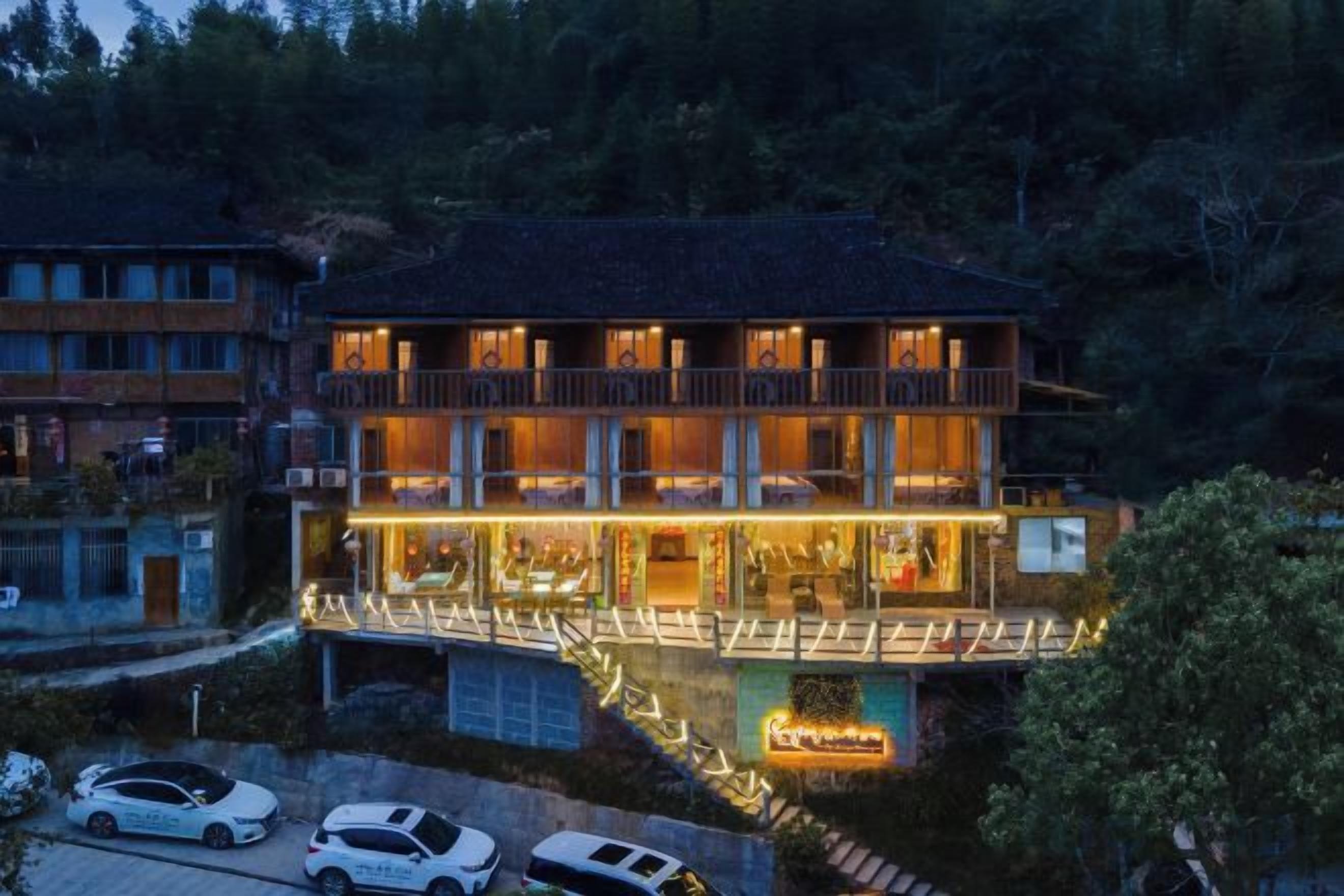 A multi-story hotel with a balcony on the ground floor, a covered outdoor seating area, and a parking lot with cars.