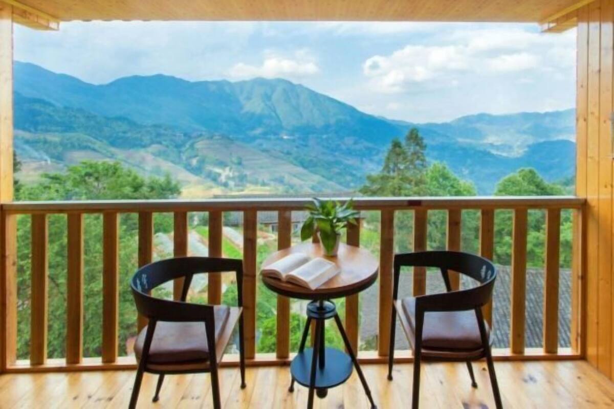 A balcony with a view of mountains, two chairs, and a table with a book and a plant.