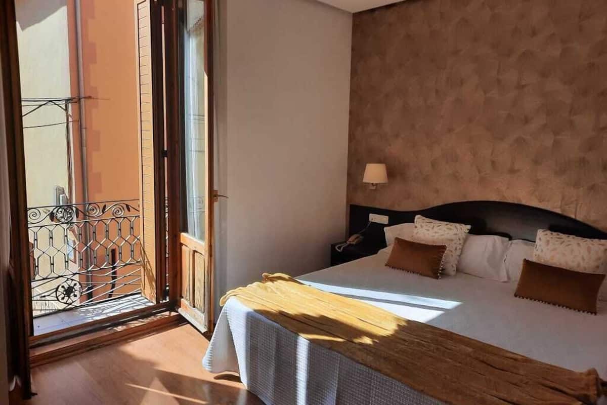 Un dormitorio con cama, mesita de noche, lámpara y vistas al balcón exterior a través de una puerta.